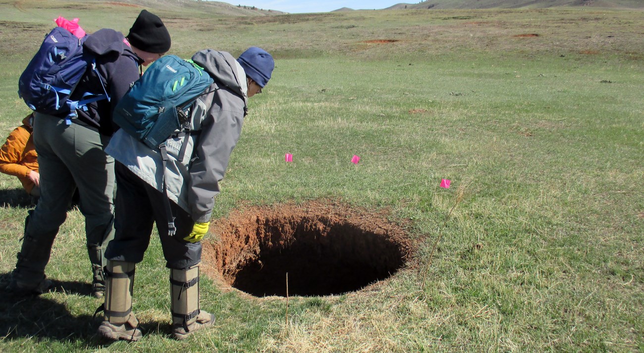 Two people with backpacks and snake gaiters peer into a large hole while surveying a grassy plain. A third person kneels nearby.