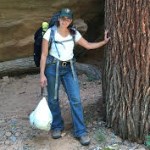 A woman in green ball cap and hiking backpack stands with hand on a tree.