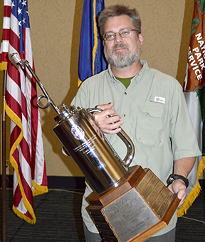 Fenn Wimberly holds a driptorch trophy in front of flags.