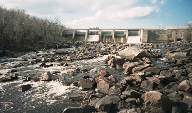 Low water flow over rocky river bed with hydro dam in background.