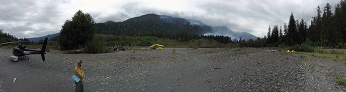 A helicopter sits on a gravel bar with a firefighter nearby and tents in the distance.