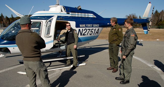 Maine Forest Service Three men stand and listen to a man pointing at something in a helicopter.