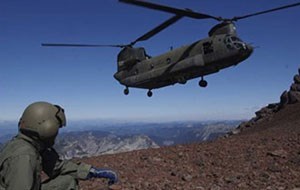 A person in a green flight suit hunches on the ground while a helicopter takes off nearby in a rocky mountainous area.