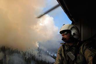 A woman in a flight helmet looks out the door of a helicopter on mountain filled with smoke.