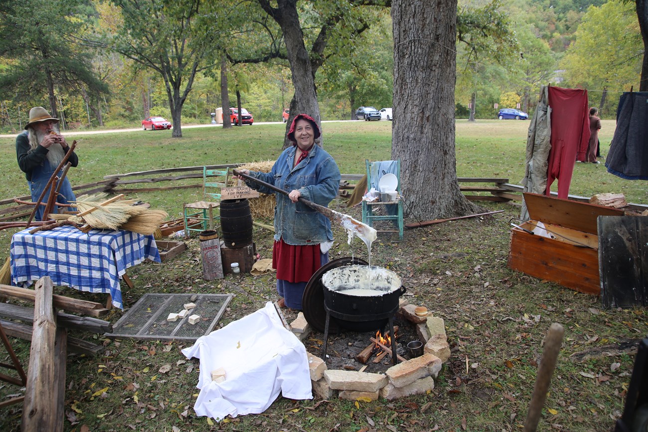 A woman holds a molten scoop of soap above a large pot.