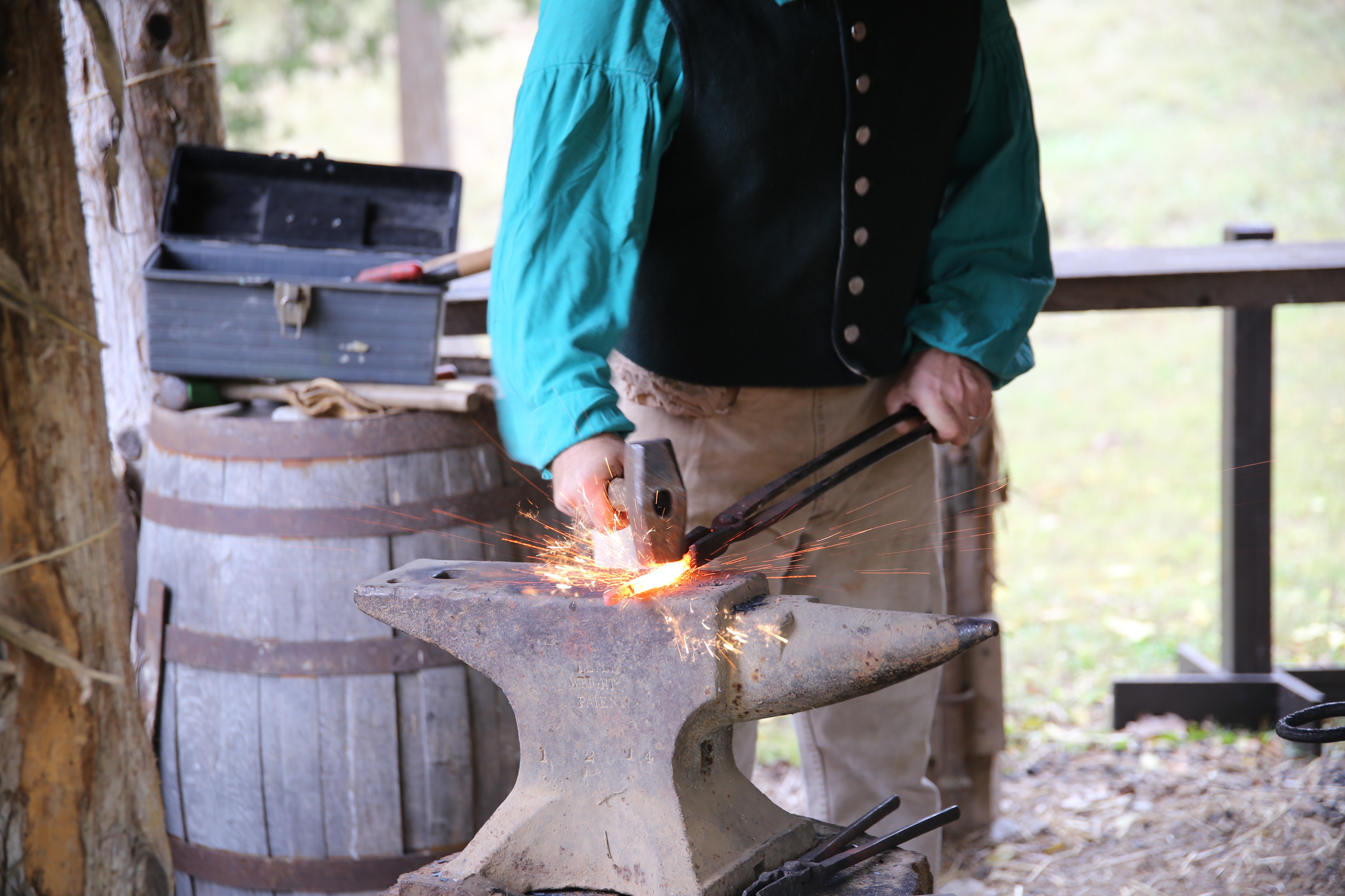 A man in a green shirt and black vest strikes hot metal with a hammer, sending sparks in all directions.