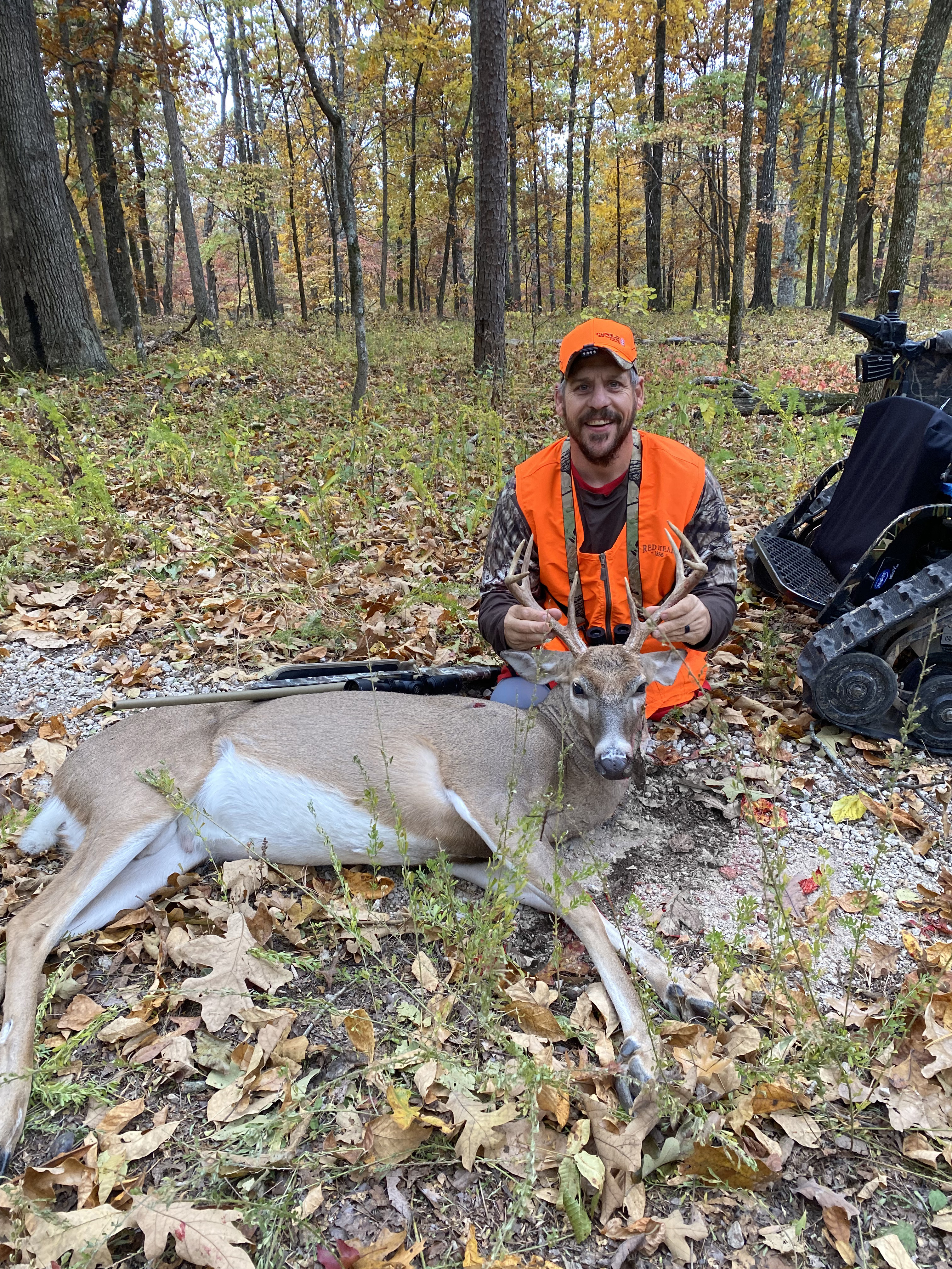 A man in a hunter orange outfit smiles at the camera. A freshly-hunted deer lies in front of him.