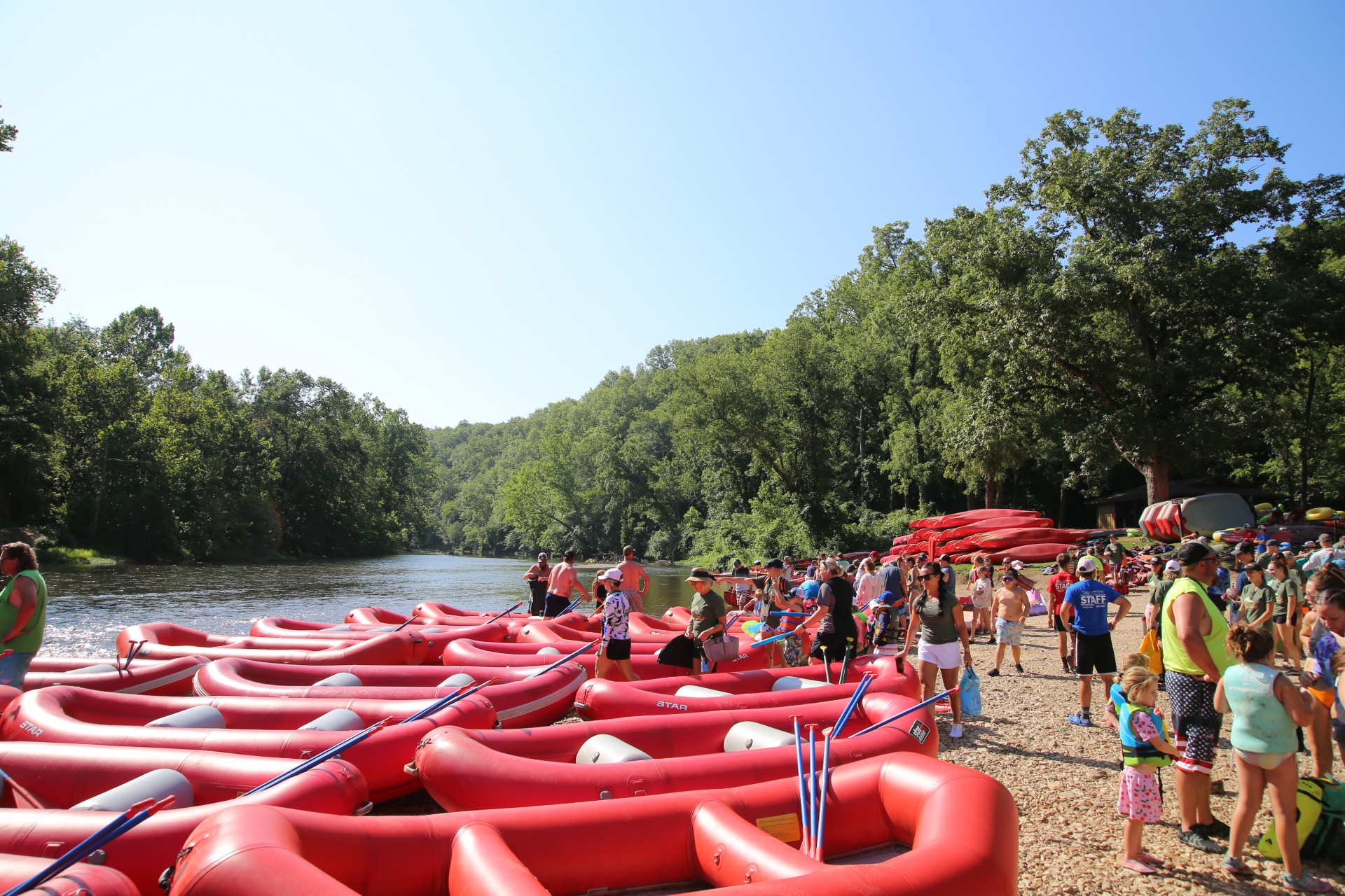 A busy gravel bar by the river. Red rafts and kayaks are piled up with visitors of all ages waiting to load in them and float.