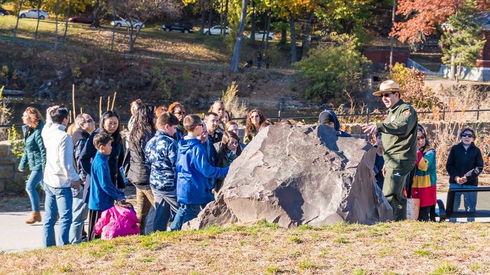 A park ranger stands beside a boulder at speaking to school children, fall foliage & the top of a waterfall visible in the background