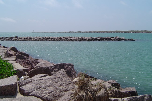 Large rocks piled into a line bordering the waters edge along two sides of a channel filled with blue-green water.