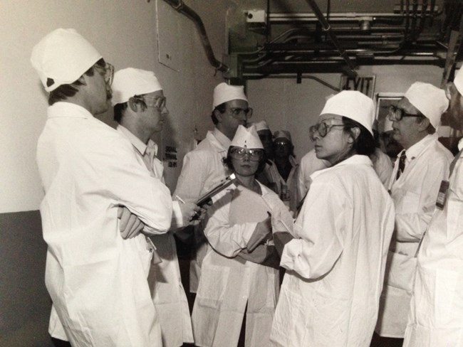 An older photo of Karen Dorn Steele in white attire standing in the middle of five journalists inside the PUREX Plant.