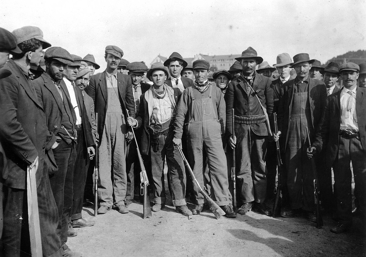 25 United Mine Workers pose for a picture outside. They are dressed in civilian clothes, like overalls, and are shown holding rifles and wearing ammunition belts.