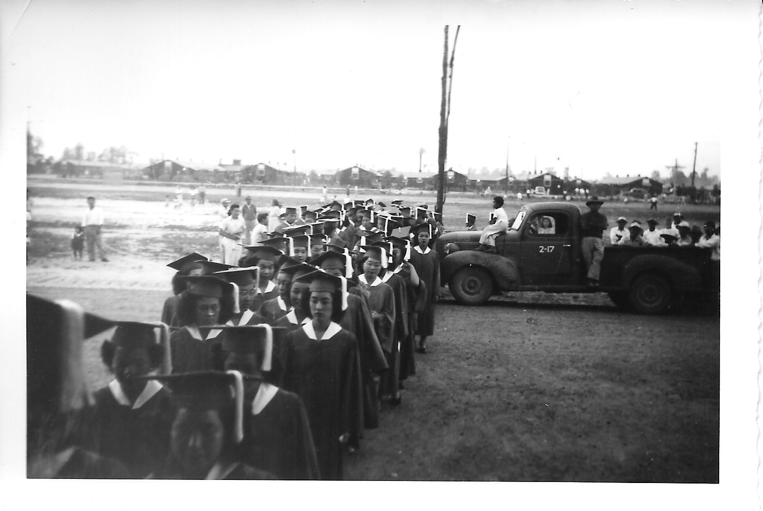 Graduates of Rohwer High School line up outside in their caps and gowns while people look on.