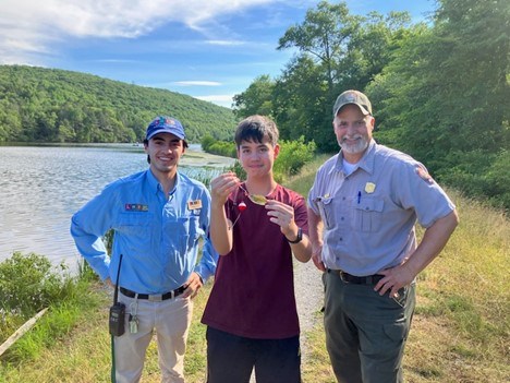 Visitor Fishing Three people stand in front of a lake with forested hills in the background. In the middle the individual is holding a fish. The two individuals on the outside are in a Latino Heritage Internship Program uniform and a Park Ranger uniform.