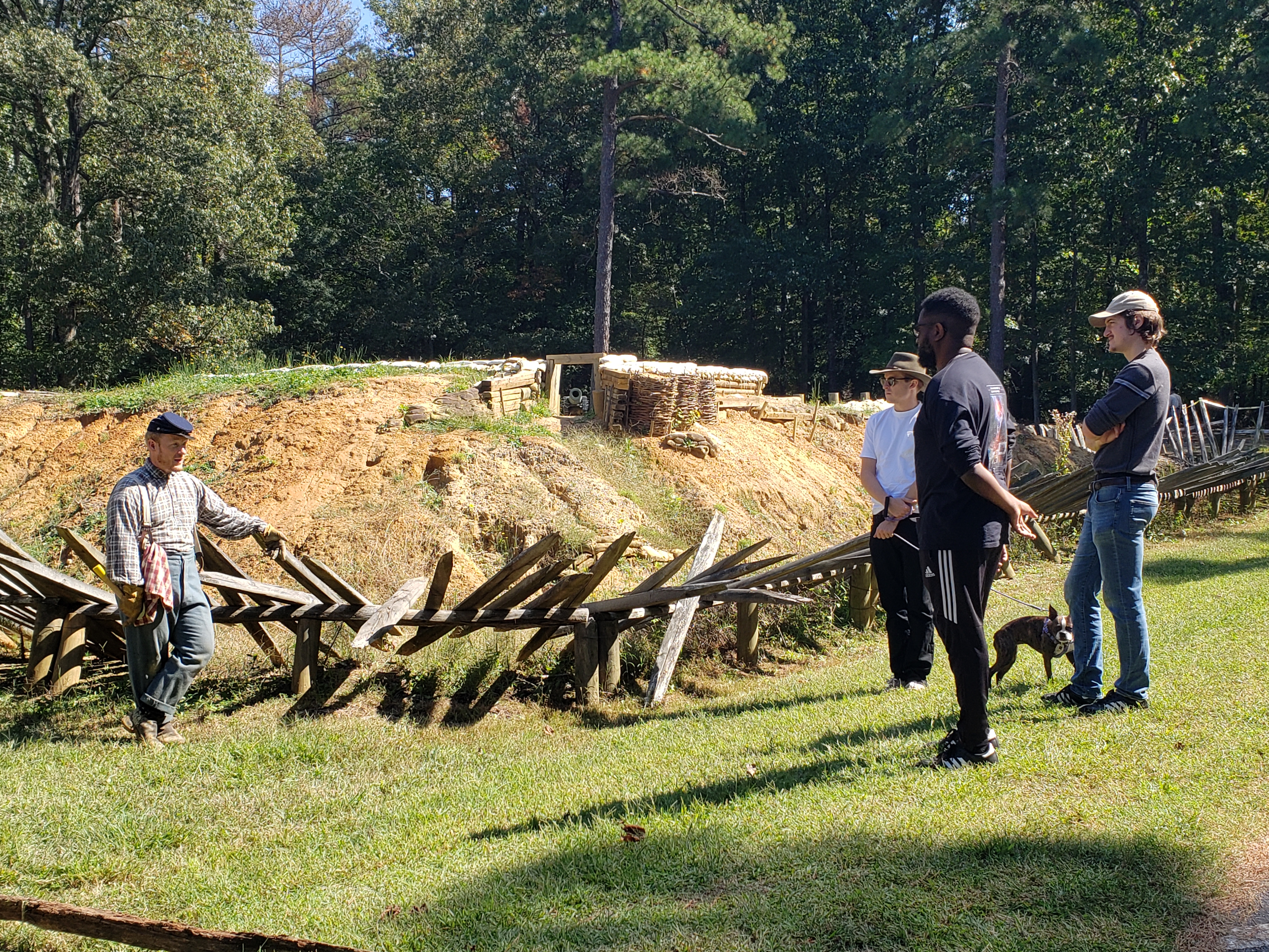 A white male in dressed as a Civil War soldier speaks to three people standing next to a reconstructed fortification.