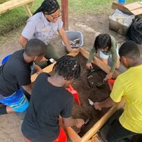 Four children with brown skin dig in a box of dirt with archeology tools.