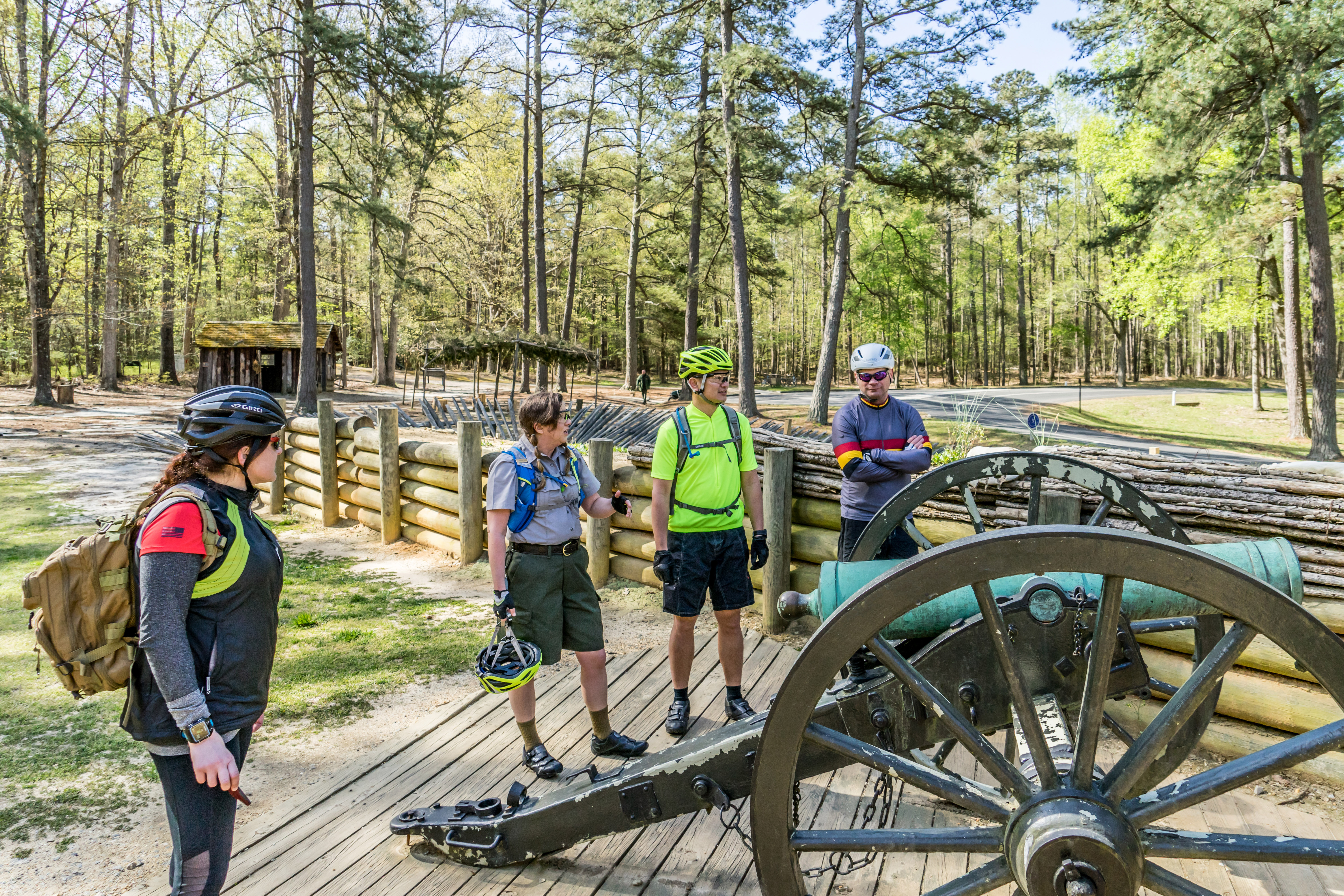 One Ranger and three visitors wearing bike helmets and backpacks stand around a green patina cannon.