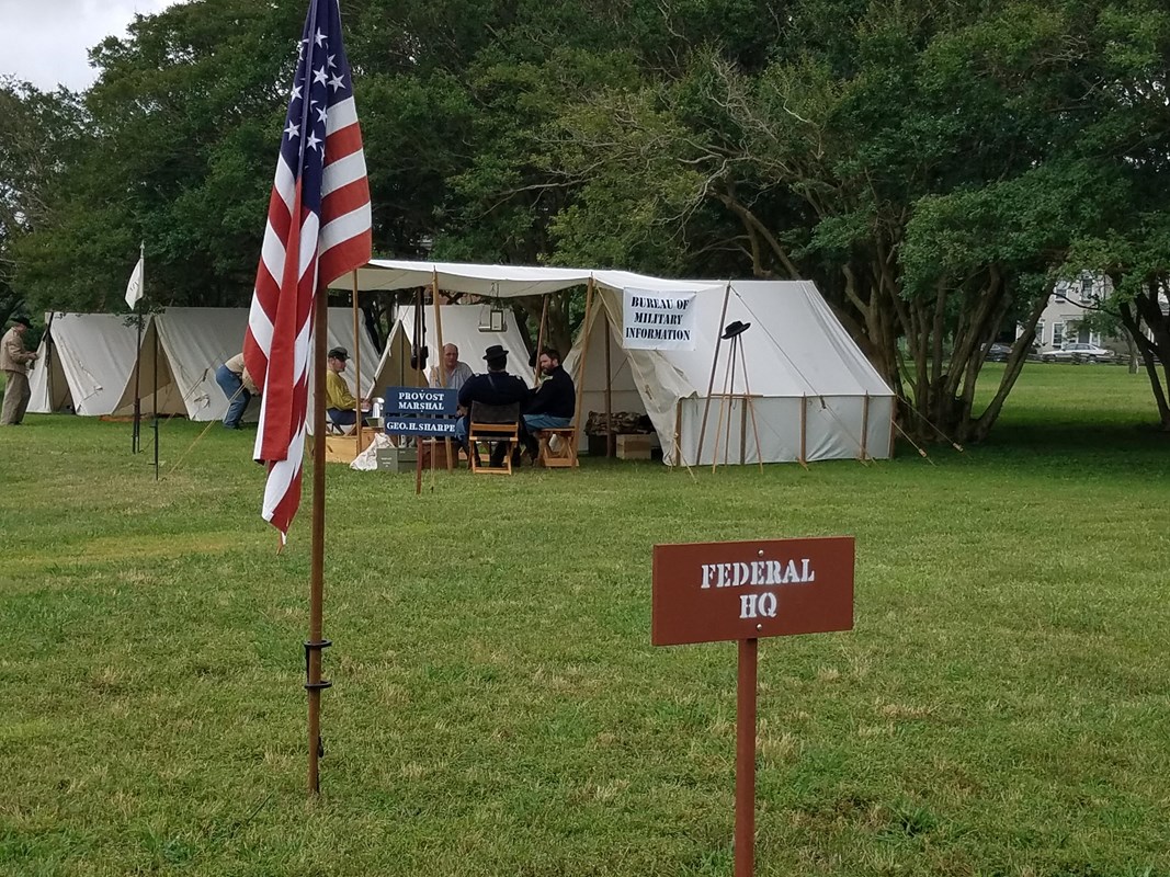 A white canvas tent next to trees with a sign that says Federal HQ. In front is the U.S. Flag on a pole.