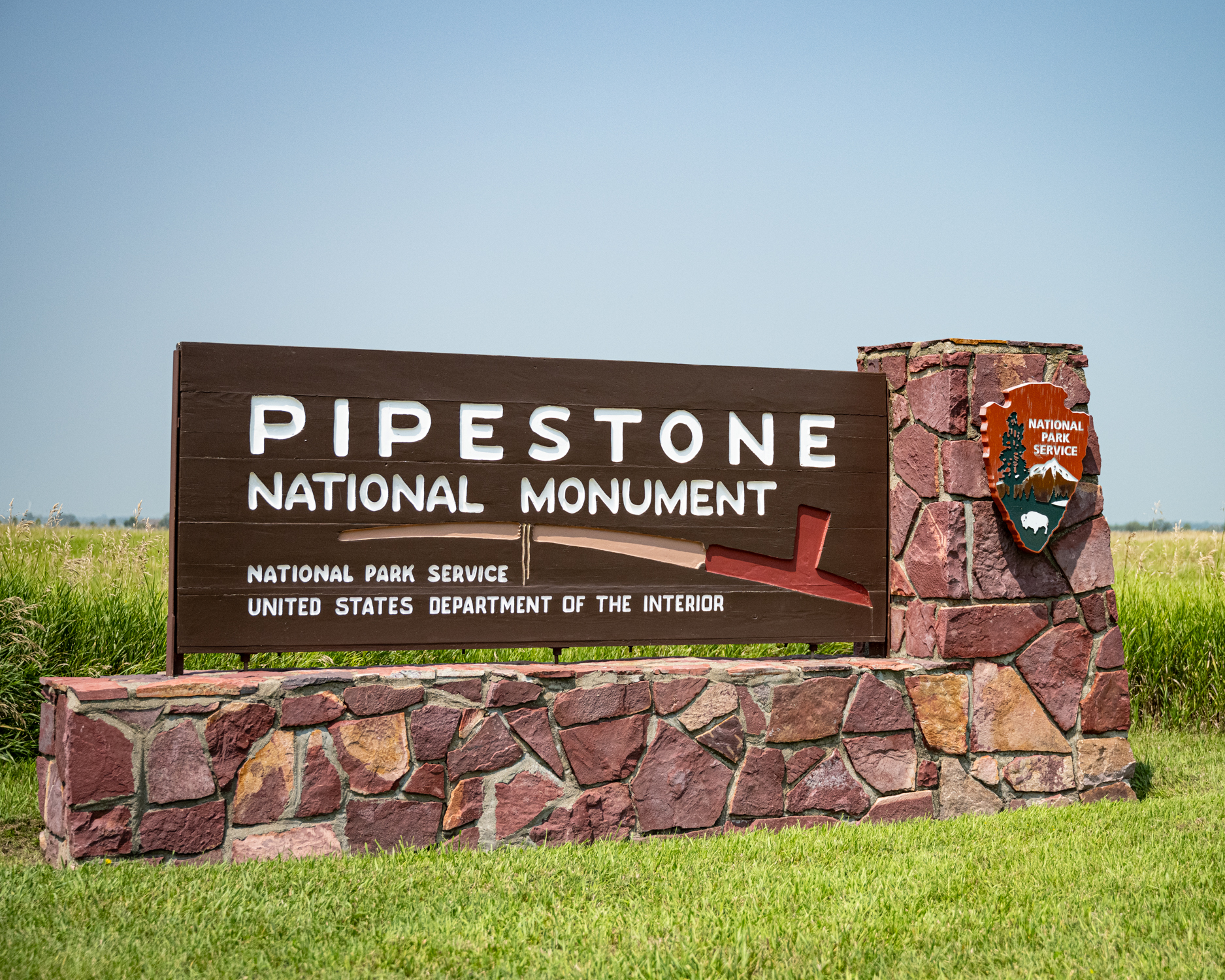 Image shows the entrance sign to Pipestone National Monument. It is a large brown sign with text reading: Pipestone National Monument, National Park Service, United States Department of the Interior. Across the center of the sign is a stylized image of a