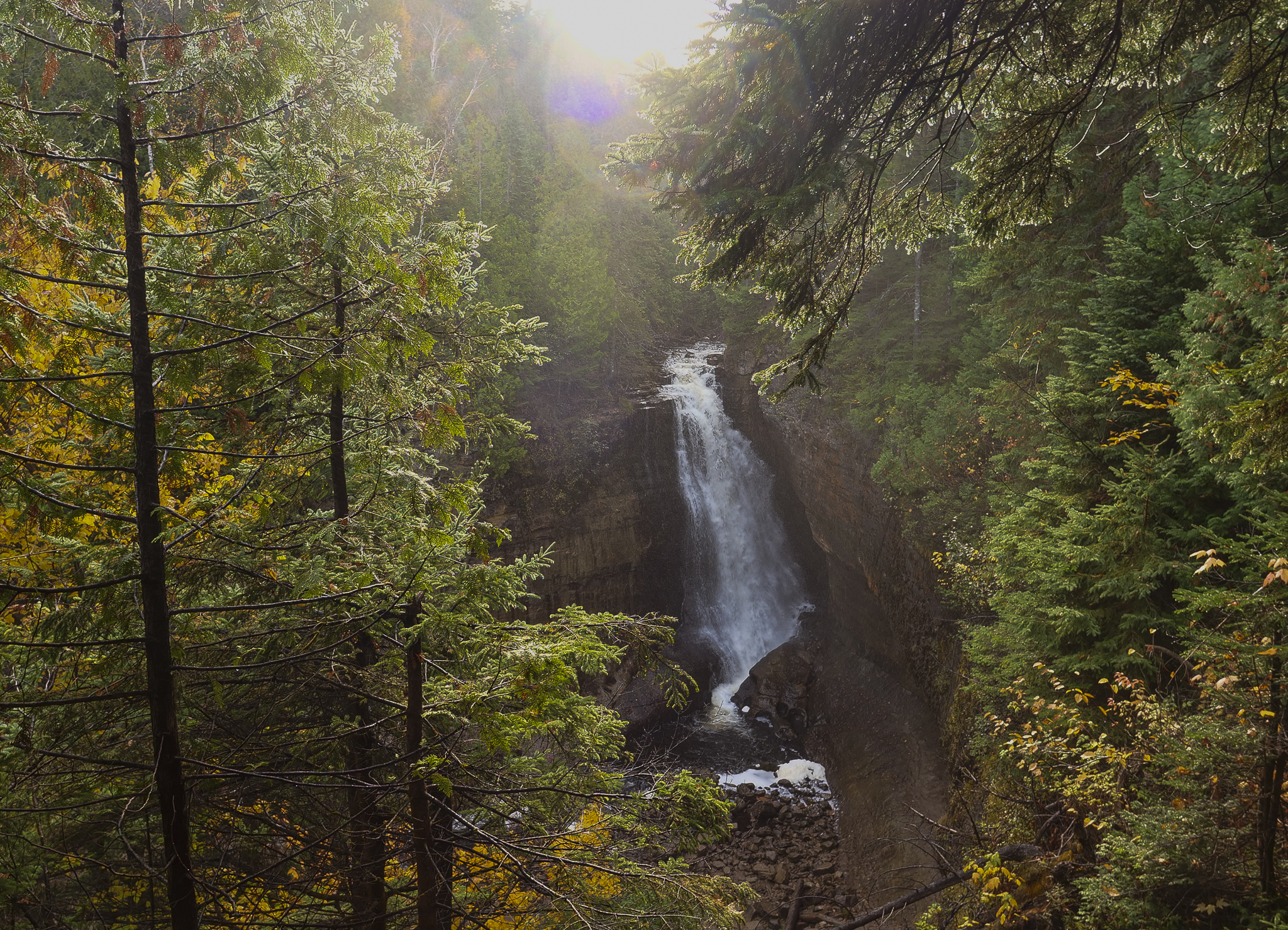 A large, powerful waterfall plunges off a sandstone cliff, surrounded by yellow and green foliage