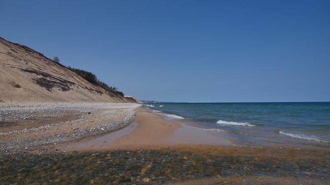 A pebble and sand beach with rising sand dunes in the background.