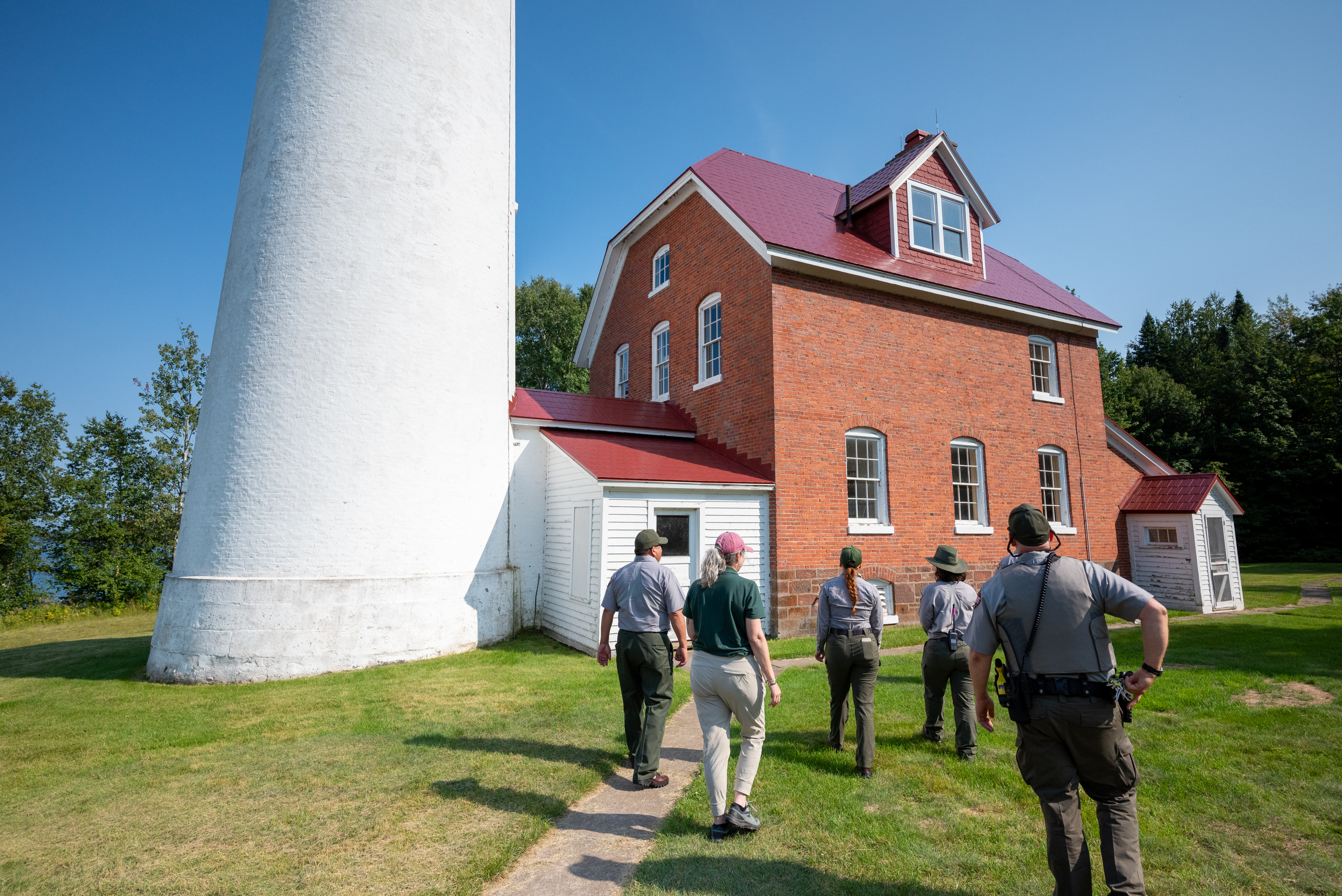 A group of people in National Park Service uniforms stand in front of a lighthouse as they walk along a path on a tour.