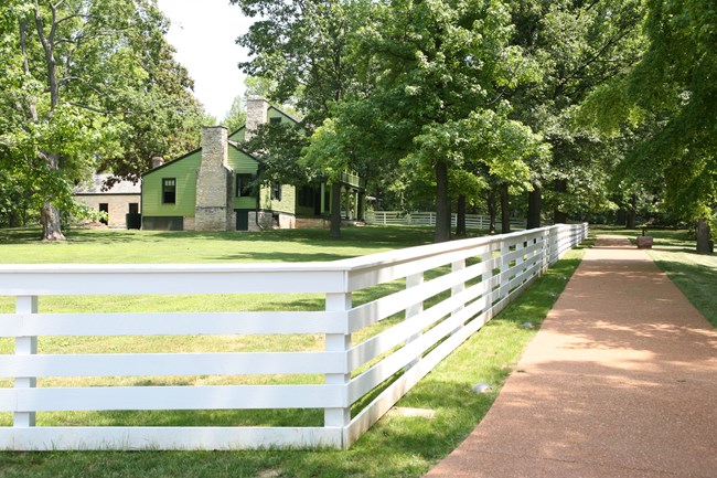 white fence on left with green house behind it. paved walkway on right running parallel to fence in front of house.