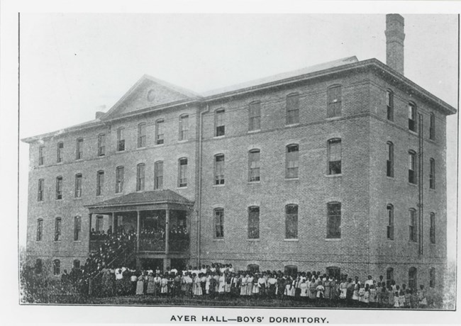 Large building with large number of people standing on front porch and around the school