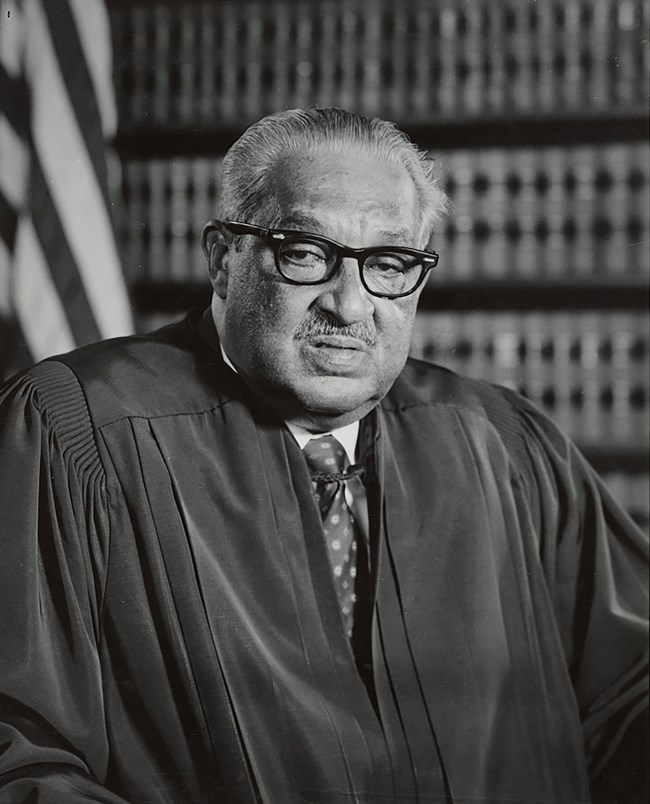 An older African American male with glasses in a judicial robe. Books and a flag behind him.