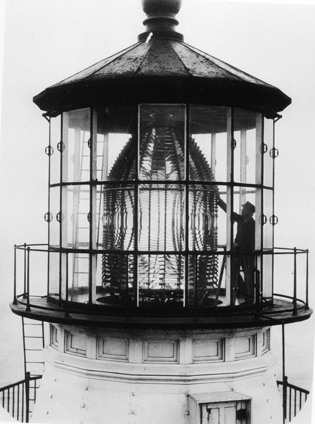 A black and white photograph of a lighthouse keeper inside the upper, glassed-in story of a lighthouse cleaning a bee-hive-shaped Fresnel lens.