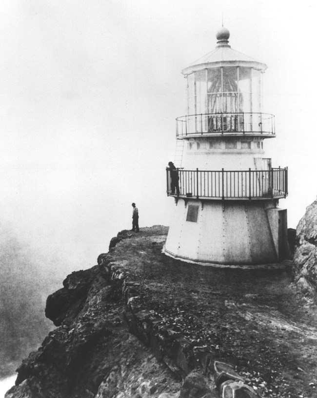 A black and white photograph of a short, white, three-story-tall lighthouse perched on the edge of a cliff.