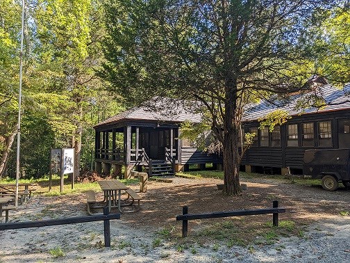 Large wooden cabin with windows, a porch, and picnic tables out front at the edge of the forest