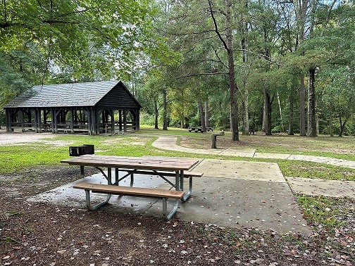 Large wooden picnic pavilion sits shaded among trees at the end of a curving paved sidewalk