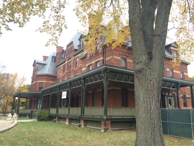 A photo of a Victorian four-story tall red brick building. A wrap around green and red porch dominates the first floor view.