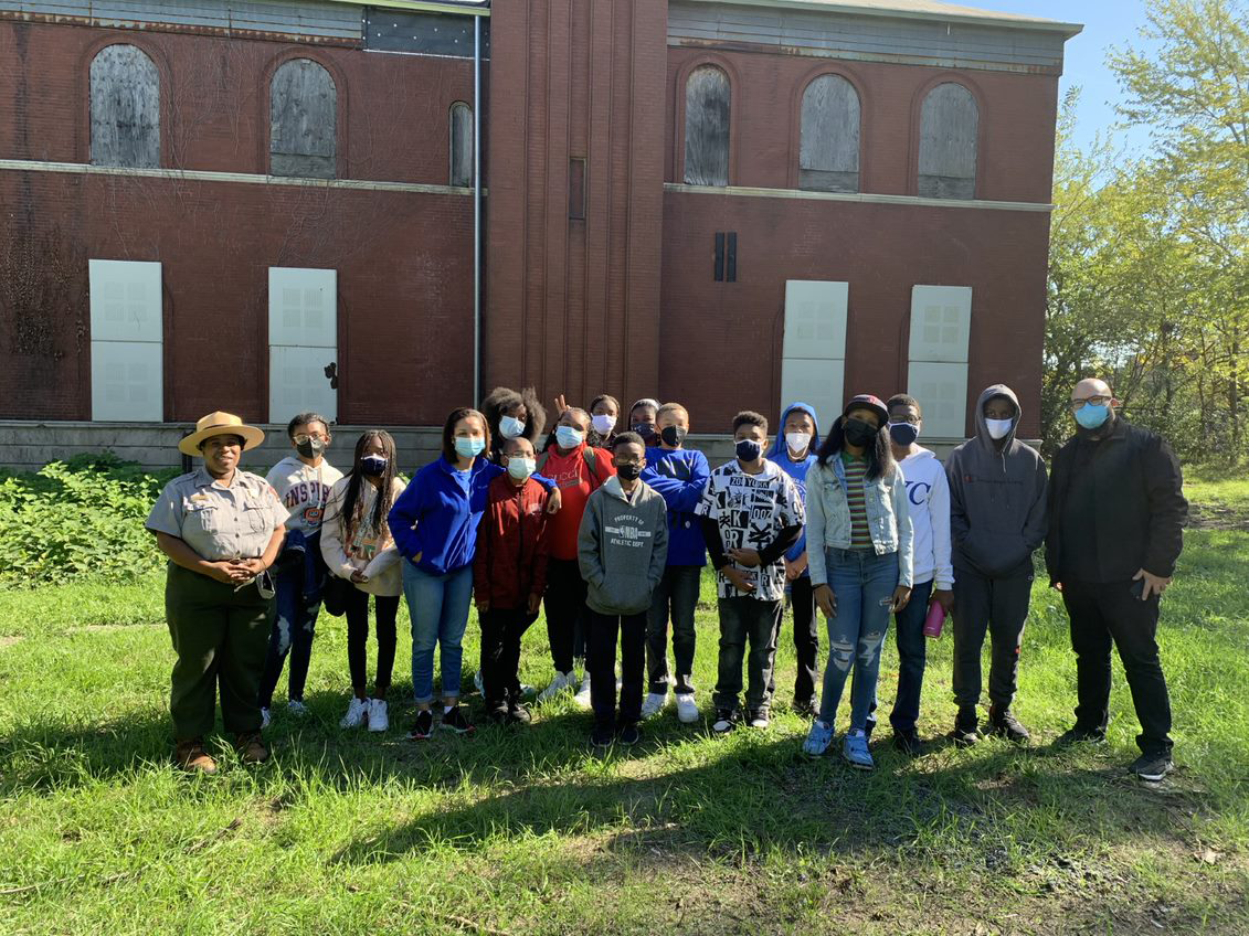 A group of students, two teachers, and a ranger pose with masks on in front of a historic brick building.