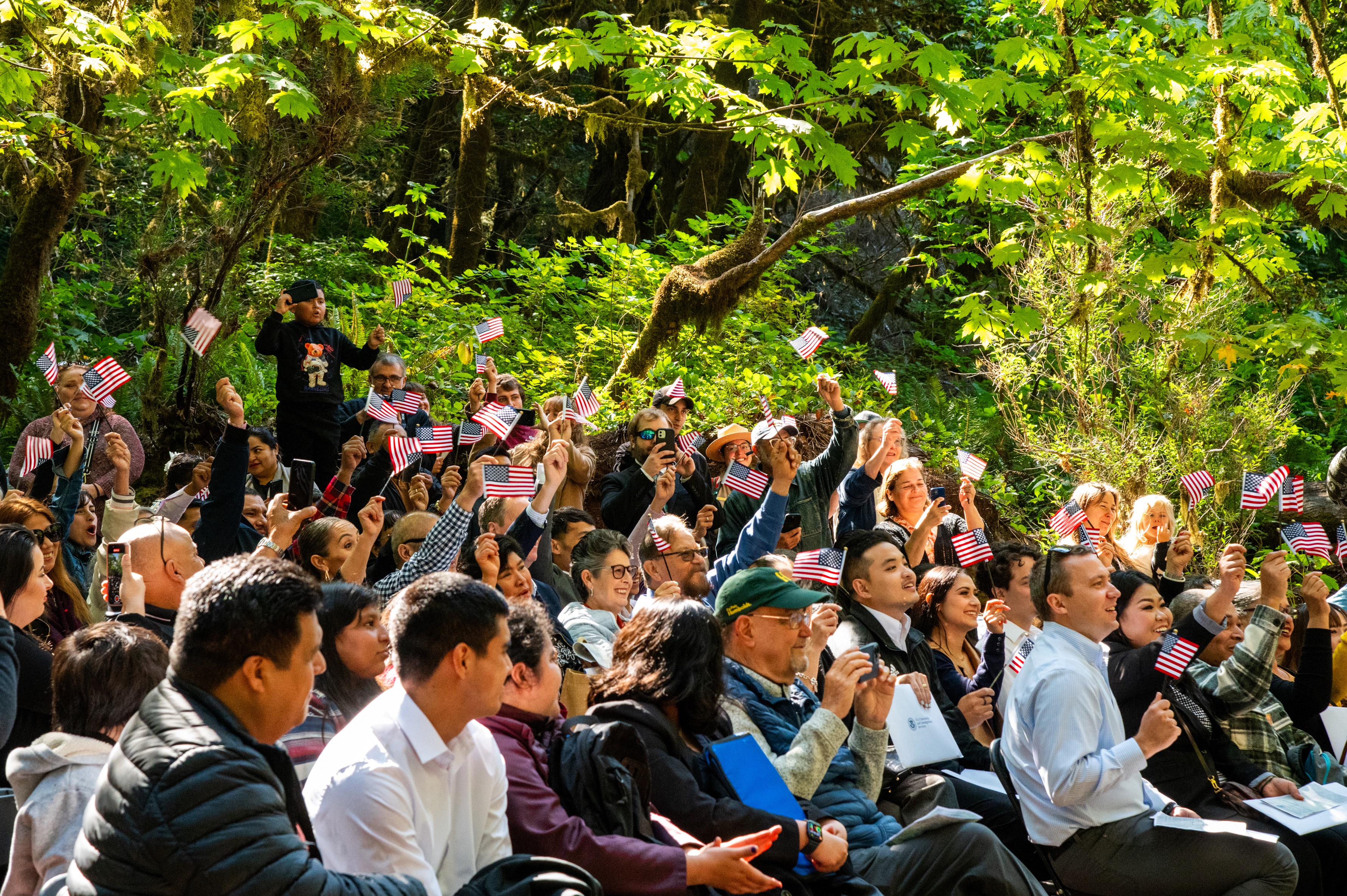 A seated group of people wave small American flags in a forest