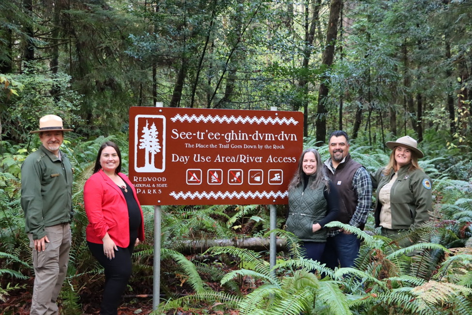 Five adults stand next to a brown sign that has Tolowa language information on it.