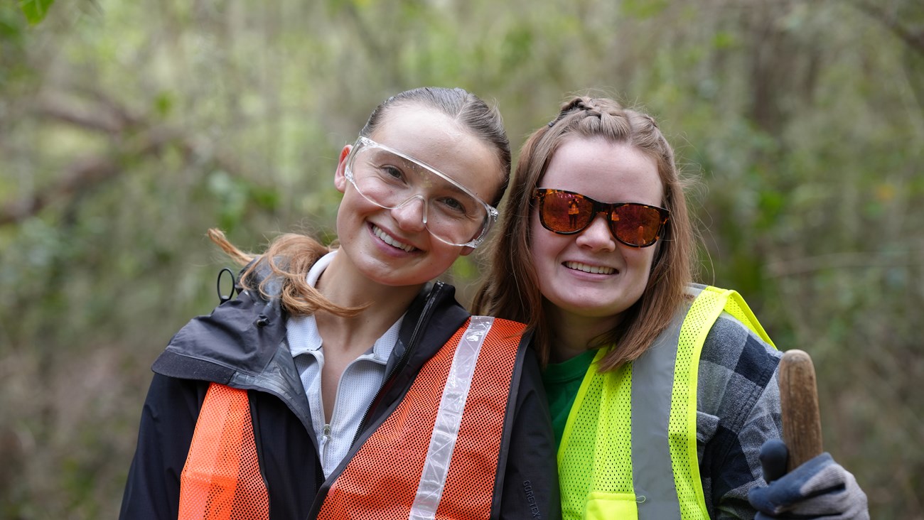 Two women wearing safety glasses and bright vests smile while they work.