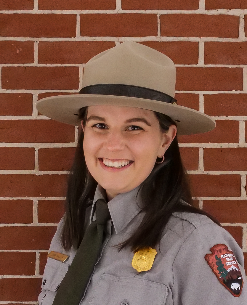 A woman wearing a flat hat and National Park Service ranger uniform smiles.