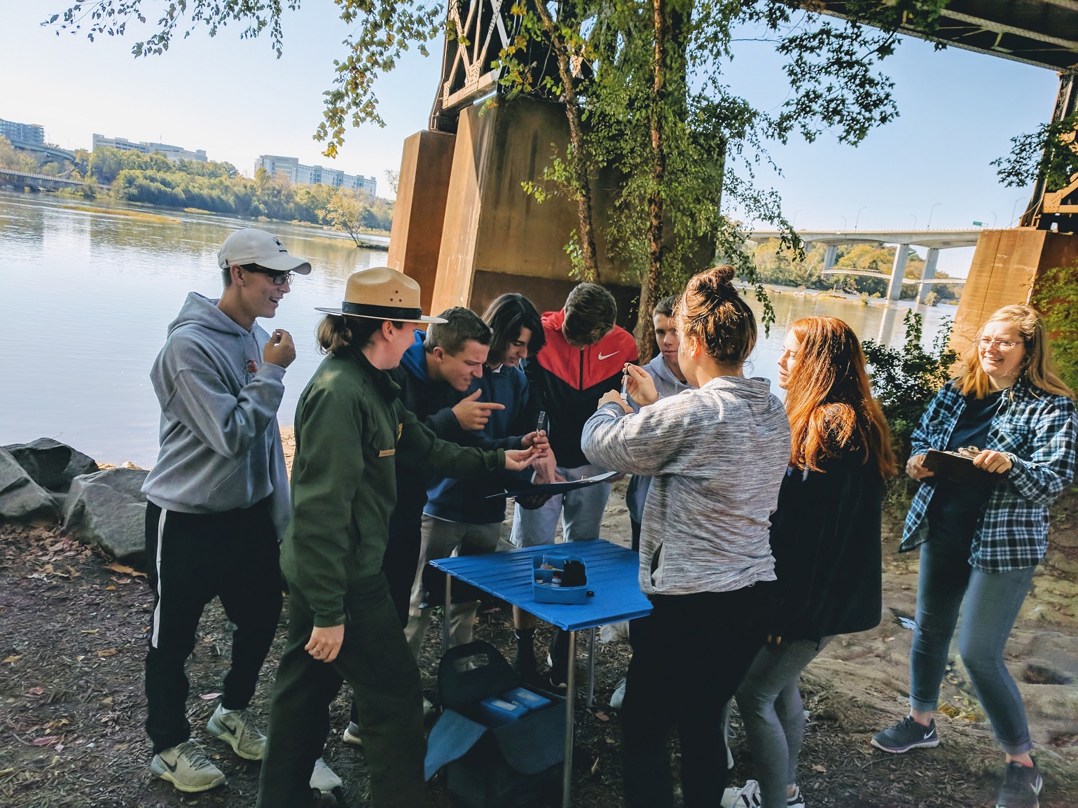 A park ranger surrounded by children at the bank of a river.