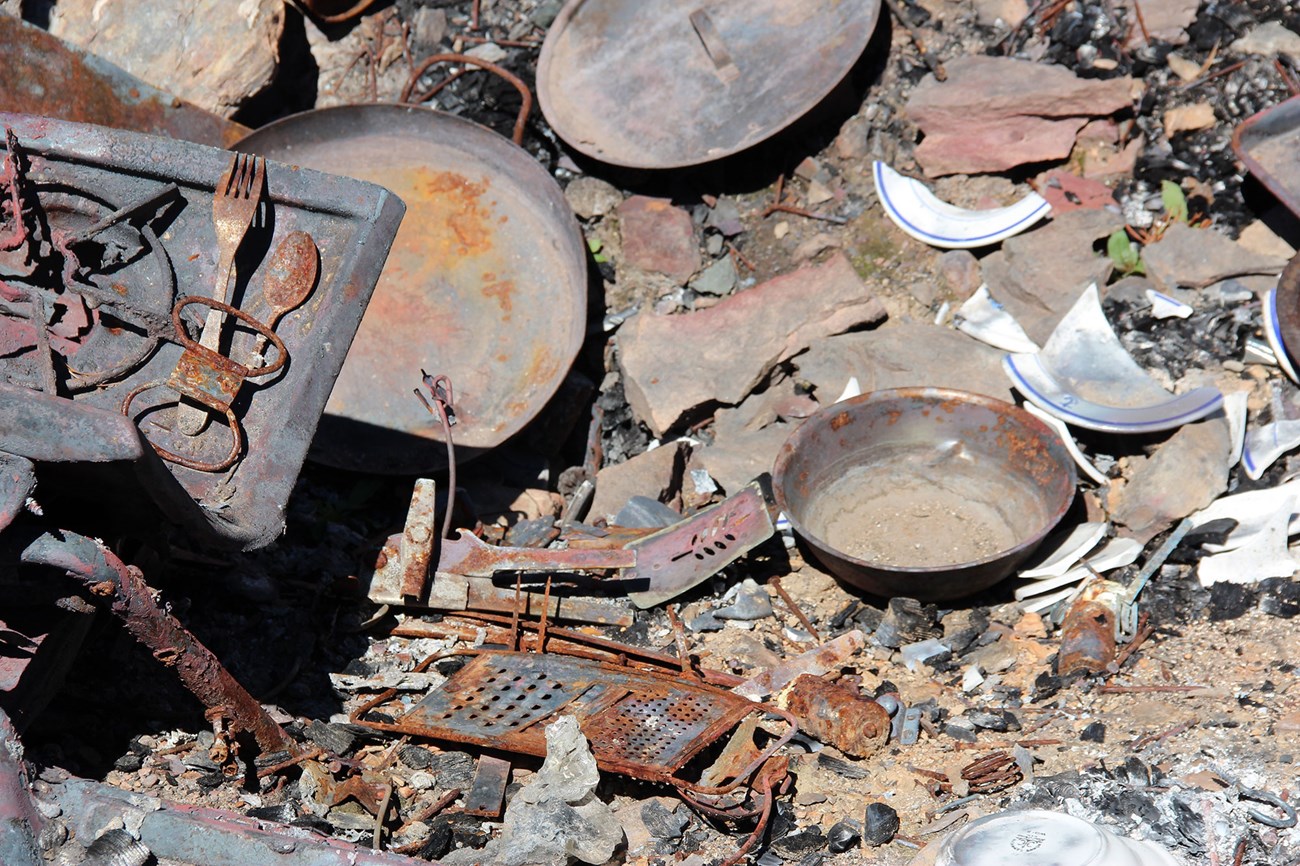 Old pans, rusted utensils, and broken plates lie in a pile. Rusted, old pans and utensils, broken plates, and a rusted, metal cheese grater lie in a pile.