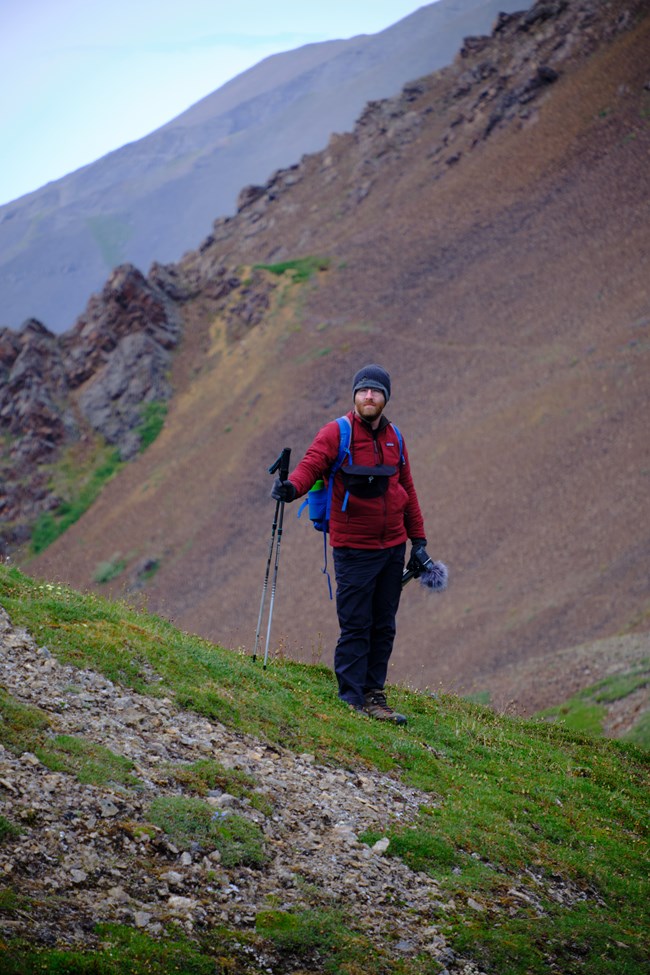 Portrait of Artist in Residence Garrison Gerard walking on a mountain trail with green grasses on the path