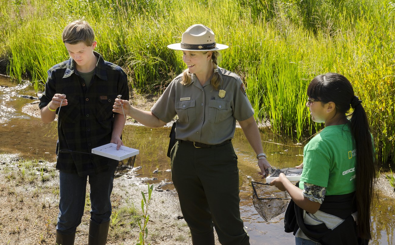 A park ranger is guiding an ecology activity, interacting with two park visitors near a river