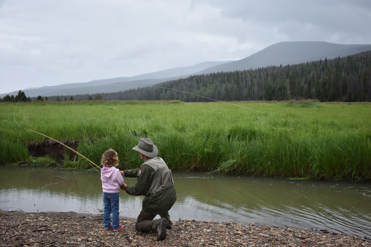 A park ranger is helping a youth learn how to cast along a riverbank.