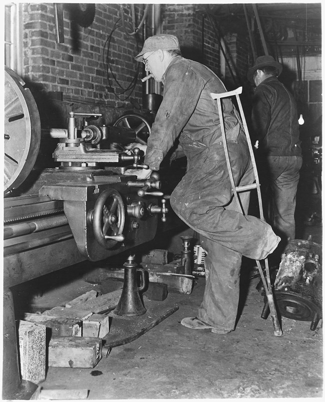 Historic photo of man without left foot, using crutch while he works in a machine shop.