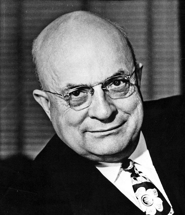 An historic photo of an older. man in a suit who is bald and wearing glasses.