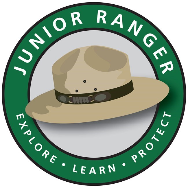 Junior Ranger logo featuring a green circular badge with a ranger hat in the center. The words "Junior Ranger" are at the top, and "Explore," "Learn," and "Protect" are at the bottom of the circle.