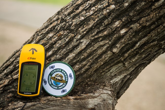 A yellow handheld GPS unit and a GPS Junior Ranger patch rest on a tree.