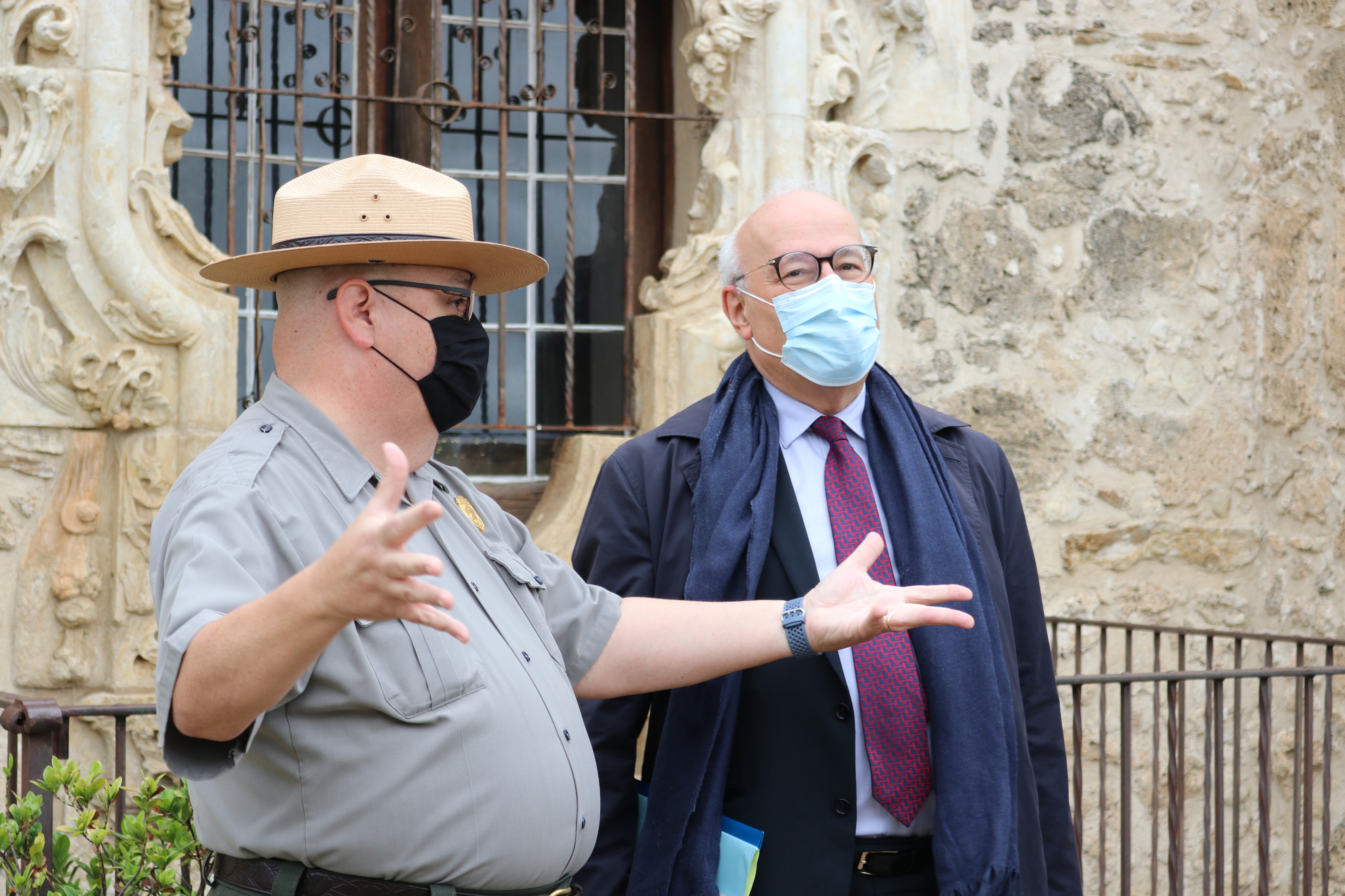 Male Park Ranger and visitor both wear masks in front of the Rose Window, a limestone carved window frame. Park Ranger gestures toward mission grounds.