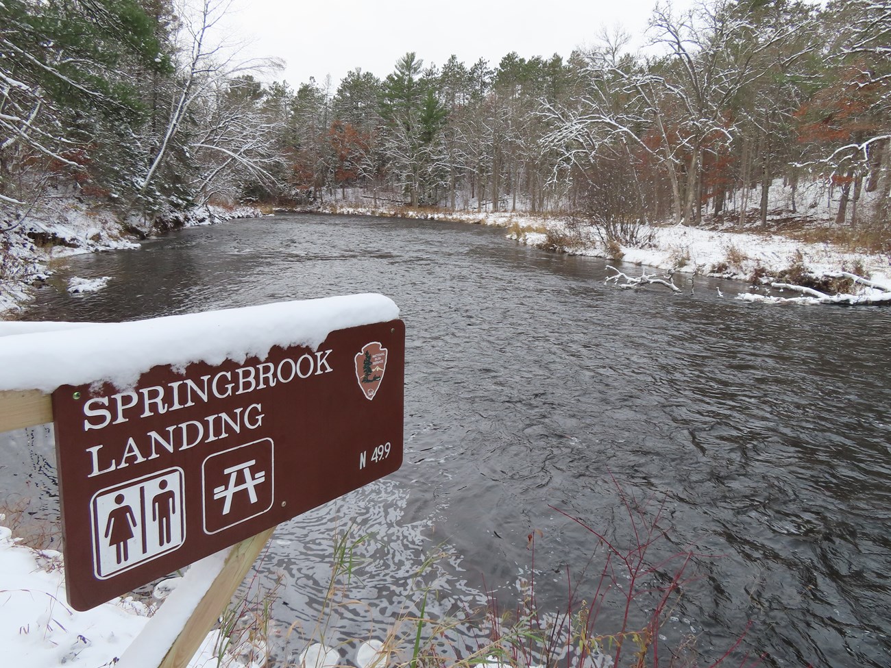 A snow-covered brown sign with white lettering reads Springbrook Landing above a flowing river with a light blanket of snow covering the stream's edges and forest.